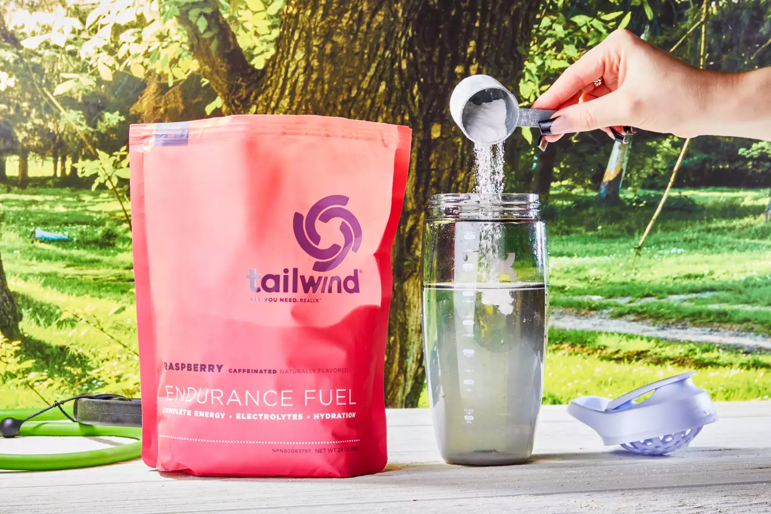 A hand pouring a scoop of Tailwind Nutrition Caffeinated Endurance Fuel into a glass of water next to the packaging