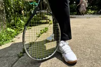 A tennis racket near Reebok Nano Court sneakers worn on concrete